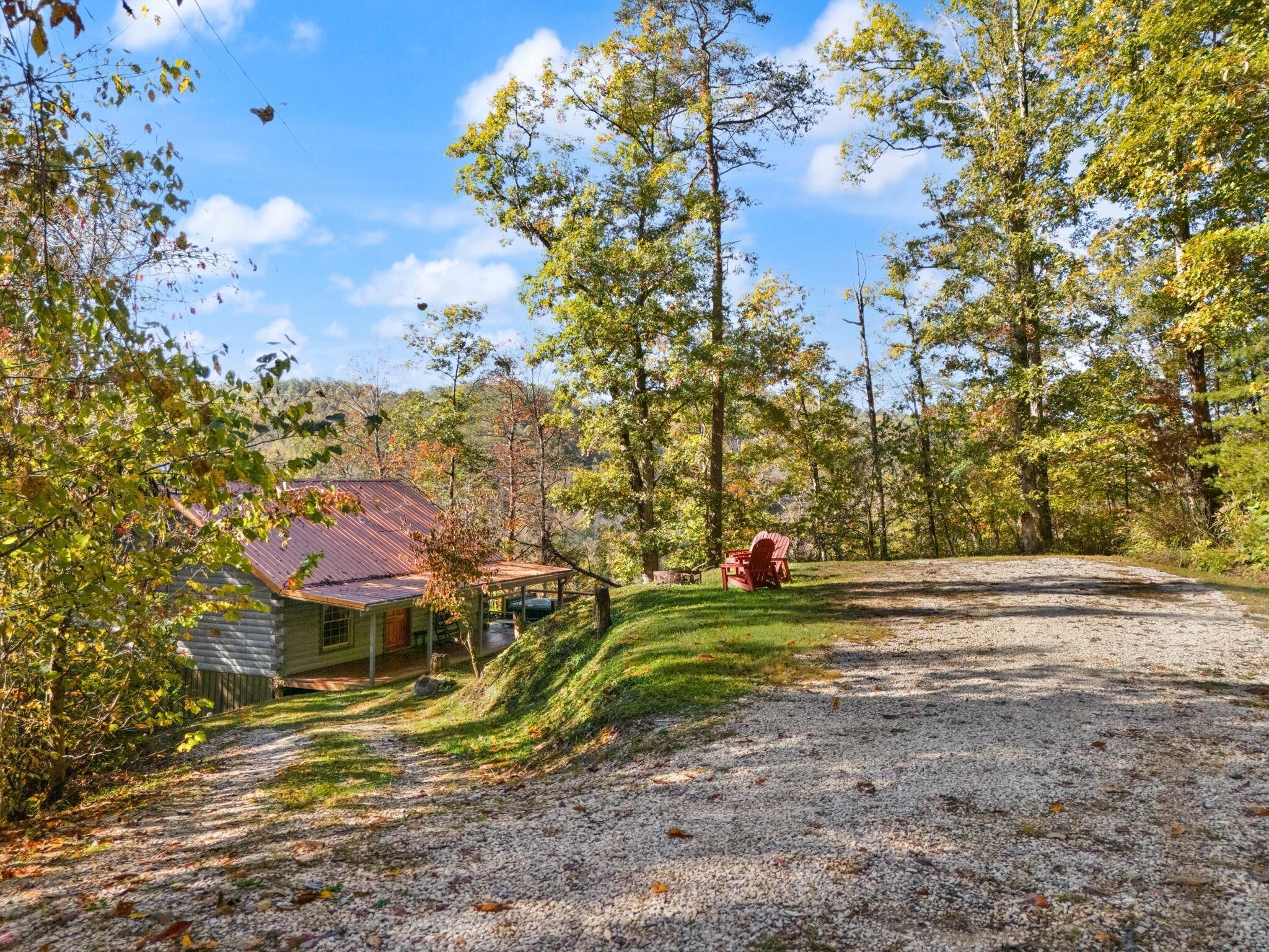 A rustic cabin with a gravel driveway, surrounded by trees and a clear sky.