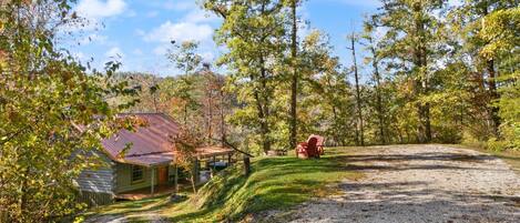 A rustic cabin with a gravel driveway, surrounded by trees and a clear sky.