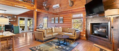 A log cabin interior with a living room, fireplace, television, and a ceiling fan.