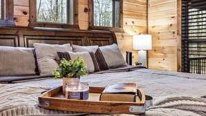 Un dormitorio de cabaña de madera acogedor con una cama, almohadas, una mesita de noche con una lámpara y una ventana.