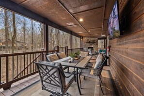 A covered outdoor dining area with a long table and chairs, large windows, and a wooden deck. - LOCATION!! Rustic/Modern Retreat~Huge Trees~Covered Deck~Hot Tub~Fire Pit~Arcade (Broken Bow)