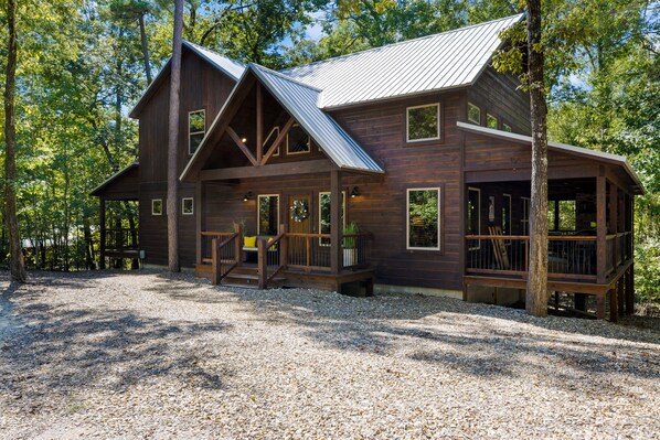 A rustic cabin with a large deck, multiple windows, and a gravel driveway surrounded by trees. - LOCATION!! Rustic/Modern Retreat~Huge Trees~Covered Deck~Hot Tub~Fire Pit~Arcade (Broken Bow)