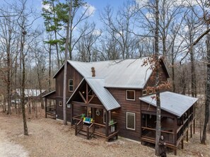 A rustic cabin with a metal roof, wooden deck, and a porch with a bench. - LOCATION!! Rustic/Modern Retreat~Huge Trees~Covered Deck~Hot Tub~Fire Pit~Arcade (Broken Bow)