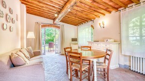 Een gezellige kamer met een houten plafond, een ronde glazen tafel met stoelen, een bank, een televisie en een keukenhoek met een raam.