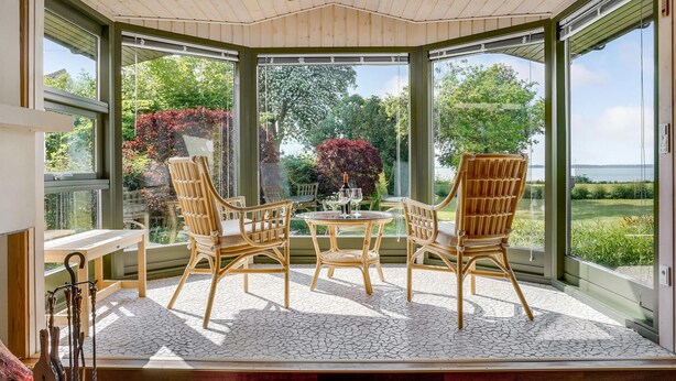A conservatory with wicker furniture, a table with glasses and a bottle, and a view of a garden and water body. - Beautiful home in Faxe with kitchen (Faxe)