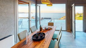 A dining area with a wooden table, chairs, and a view of the sea through large windows.