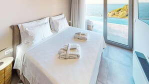 A hotel room with a large bed, two pillows, a nightstand, a window with a view of the sea, and a towel on the bed.