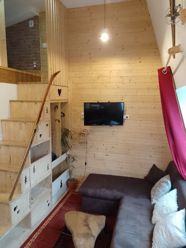 A living room with a wooden staircase, a flat-screen TV mounted on the wall, a sofa, and a red curtain. - Appartement Chaleureux Station le Lioran (Saint-Jacques-des-Blats)