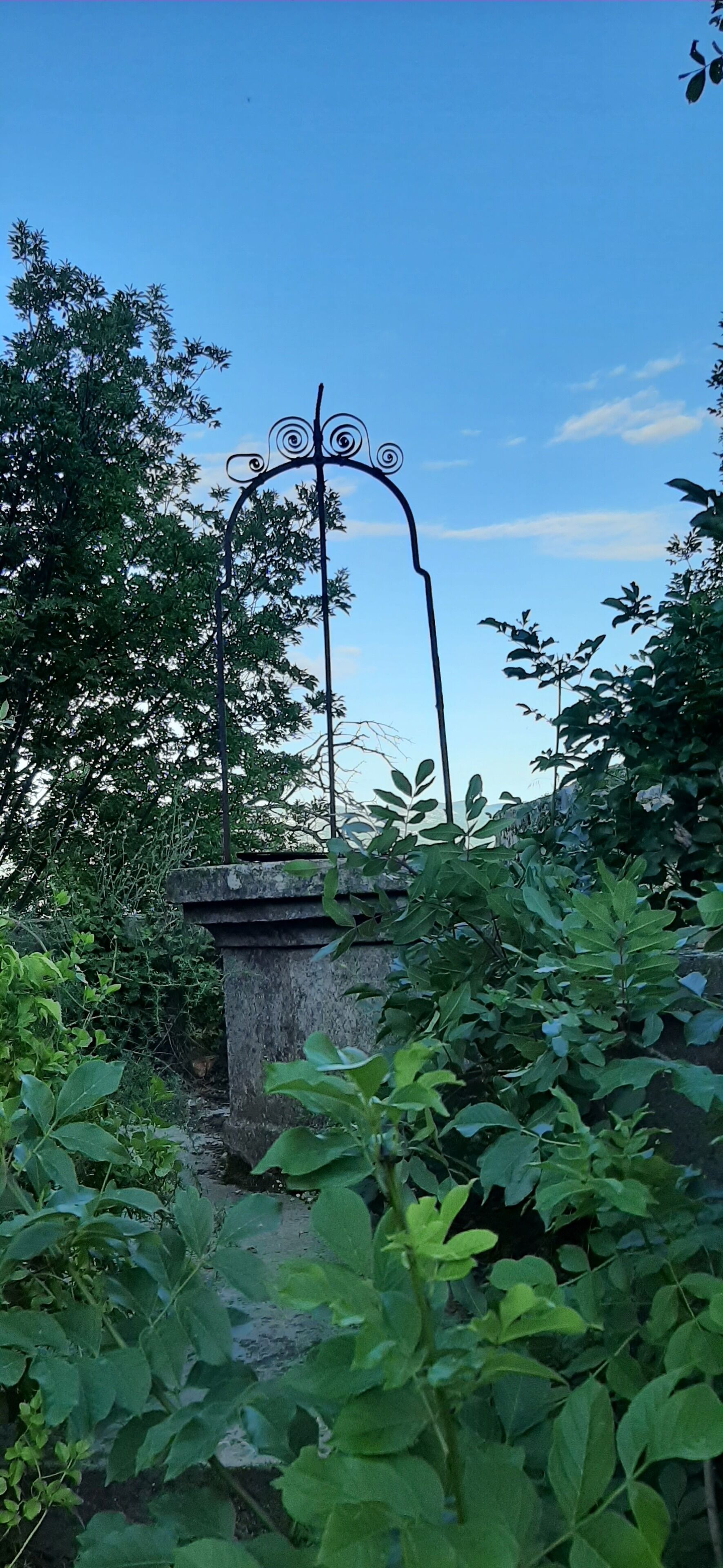 Une structure en pierre surmontée d’une arche métallique, le tout entouré d’un feuillage dense.