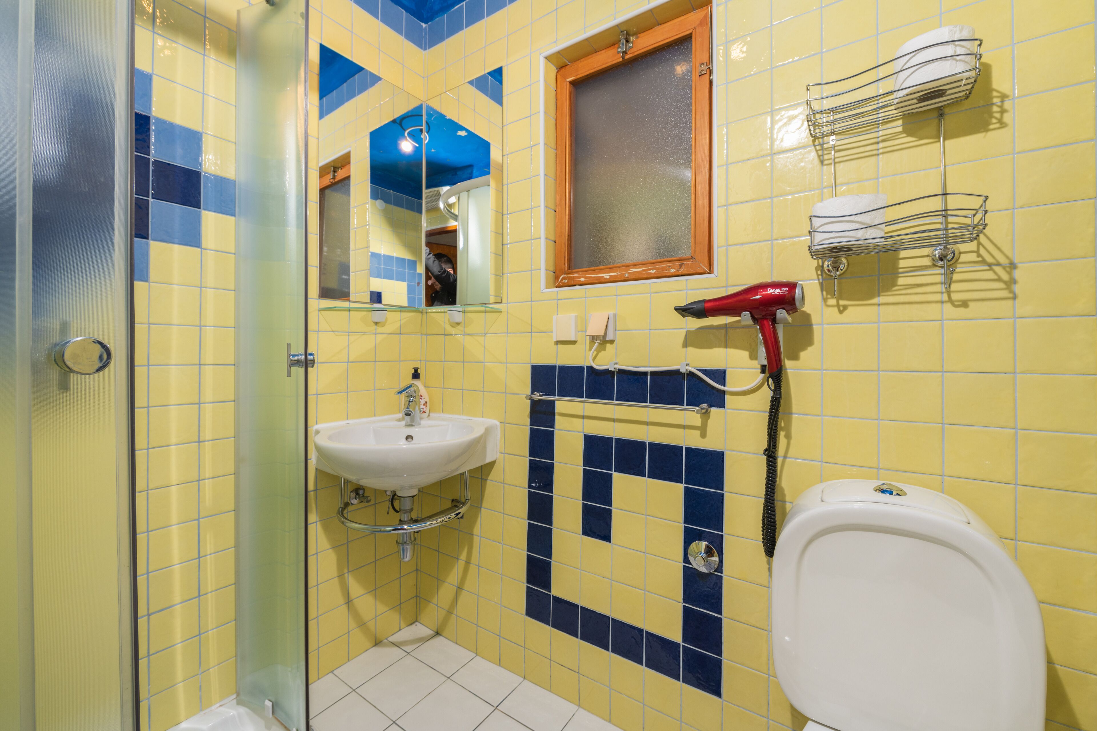A bathroom with yellow and blue tiles, a glass shower enclosure, a sink, a toilet, and a hairdryer on a rack.