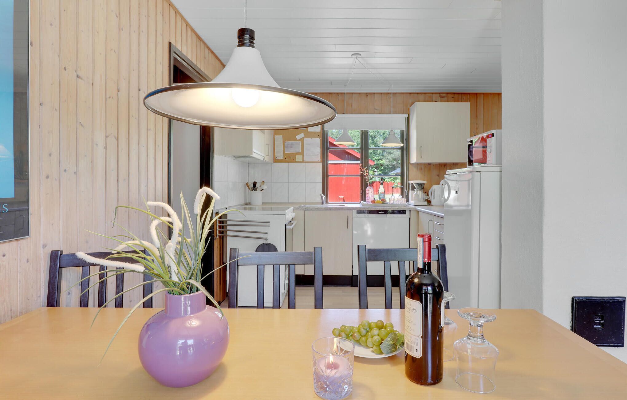 A dining area with a table set for two, featuring a purple vase with plants, a bottle of wine, and a plate of grapes.