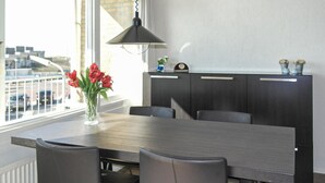 Un espace repas moderne avec une table en bois, des chaises noires, un vase de fleurs rouges et un buffet sombre agrémenté d’une horloge et d’objets décoratifs.