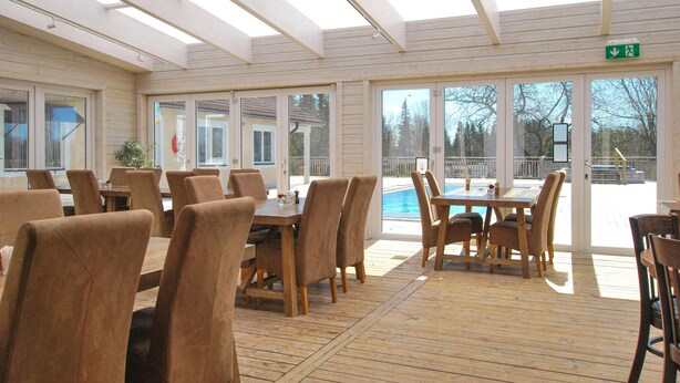 Una sala da pranzo luminosa con tavoli e sedie in legno, ampie finestre e vista su una piscina all'aperto.