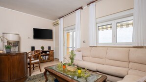 Una sala de estar con un sofá color beige, una mesa de centro de vidrio con adornos, un aparador de madera, un televisor y un ventanal con cortinas.