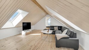 Ett modernt sovrum i loftstil med ett sluttande tak, trägolv, en svart lädersoffa, ett litet vitt bord och en platt-TV.