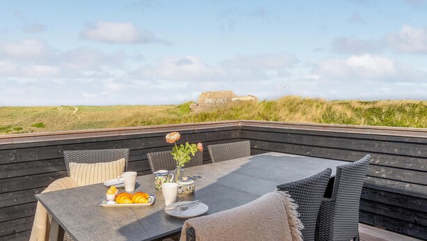 En träveranda med ett bord dukat för frukost, med utsikt över ett sanddynelandskap och en skylt med texten ”PRIVAT”.