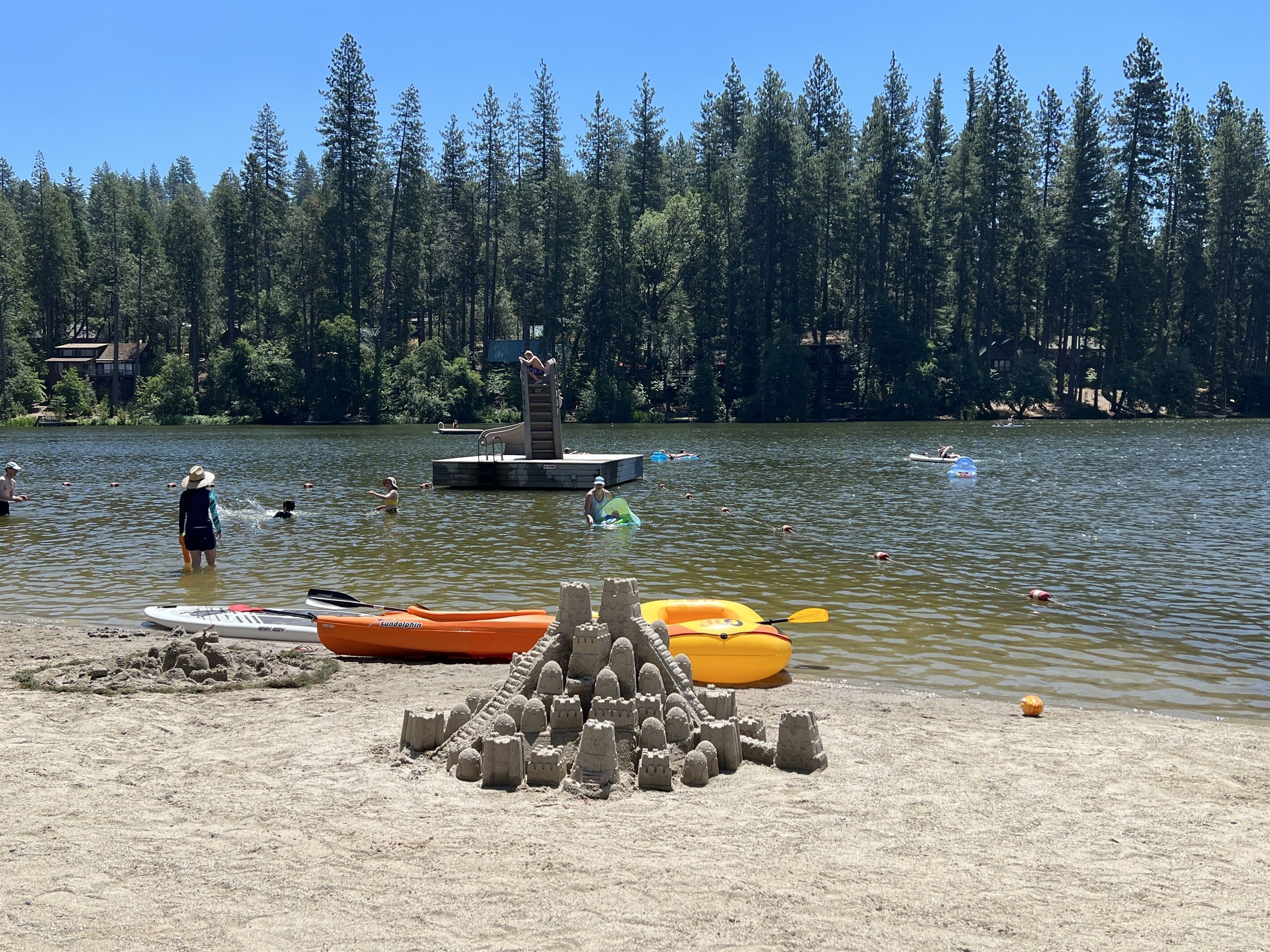 Ein sandiger Strand mit einer Sandburg, einem kleinen Steg und einem Wald im Hintergrund.