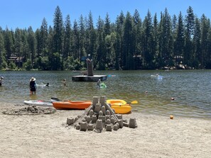 Una playa arenosa con un castillo de arena, un pequeño muelle y un bosque al fondo.
