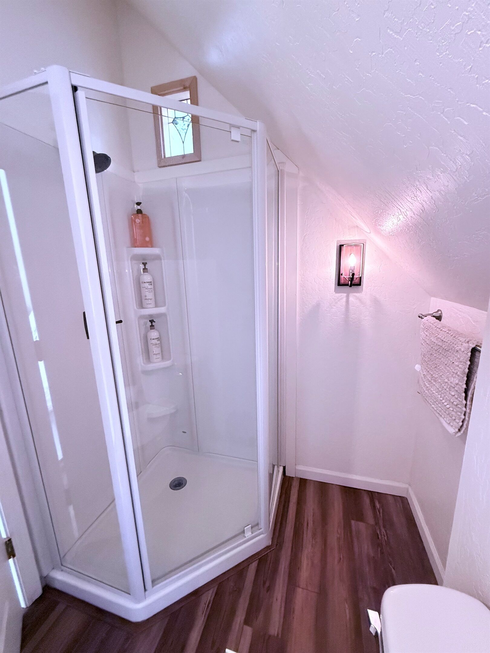 A corner shower with a glass door, a small shelf with toiletries, a towel rack, and a light fixture on the wall.