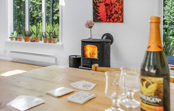 A cozy room with a wood-burning stove, a table with playing cards, a bottle of wine, and a window with potted plants.