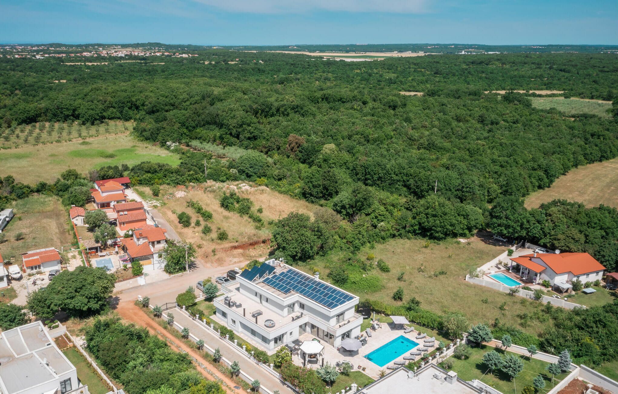 A large, modern house with a swimming pool, surrounded by dense forest and a small village in the background.