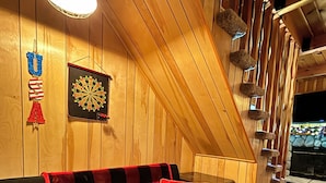 A cozy room with a wooden sloped ceiling, a black sofa with a red and black checkered blanket, a dartboard, and a fireplace. - Cozy Cabin • Near Zoo, Golf, Trails, Lake • Family & Dog Friendly • Game Room (Big Bear Lake)