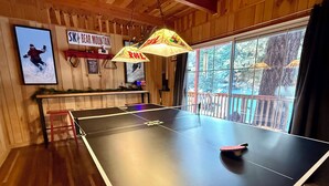 A ping pong table in a room with wooden walls, a large window with a view of trees, and a sign reading "SKI BEAR MOUNTAIN". - Cozy Cabin • Near Zoo, Golf, Trails, Lake • Family & Dog Friendly • Game Room (Big Bear Lake)