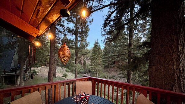 A wooden deck with a round table and chairs, a hanging lantern, and a view of a forest. - Cozy Cabin • Near Zoo, Golf, Trails, Lake • Family & Dog Friendly • Game Room (Big Bear Lake)
