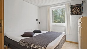 Een slaapkamer met een bed, kussens, een plaid, een lamp, een raam en een dartbord.