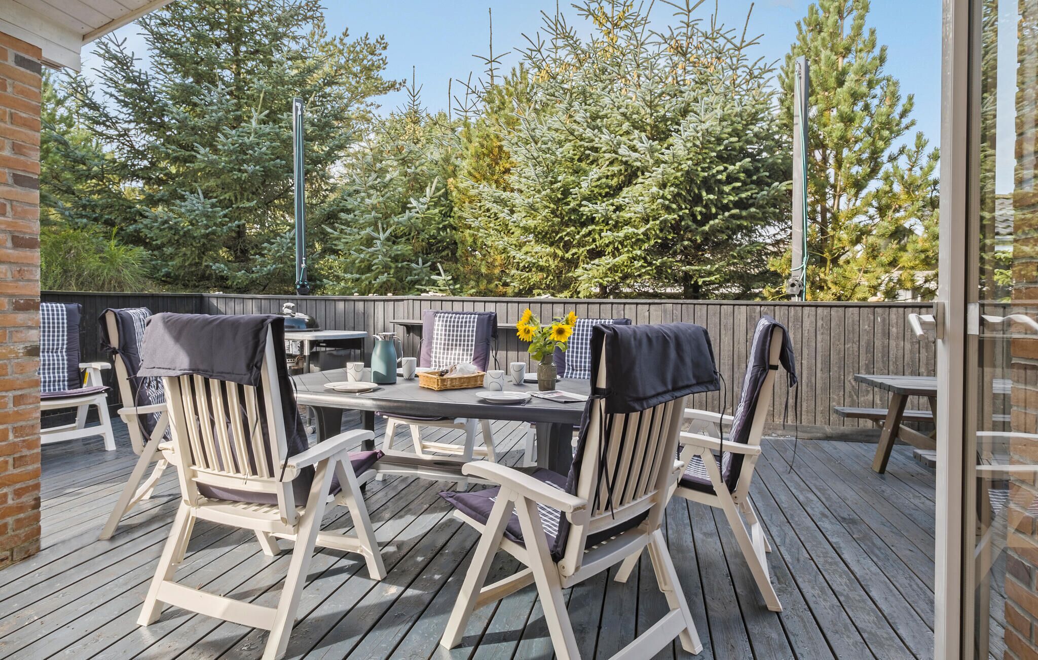 Une terrasse extérieure avec une plateforme en bois, une table et des chaises, et une vue sur des arbres et une clôture en arrière-plan.