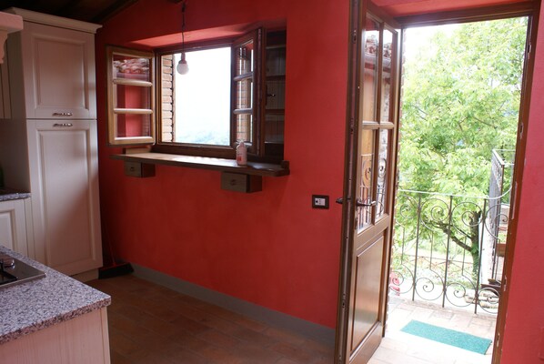 A kitchen with white cabinets, a window with a shelf, and a door leading to a balcony with a metal railing. - A Tuscan tradition, renovated in 2021 (Barga)