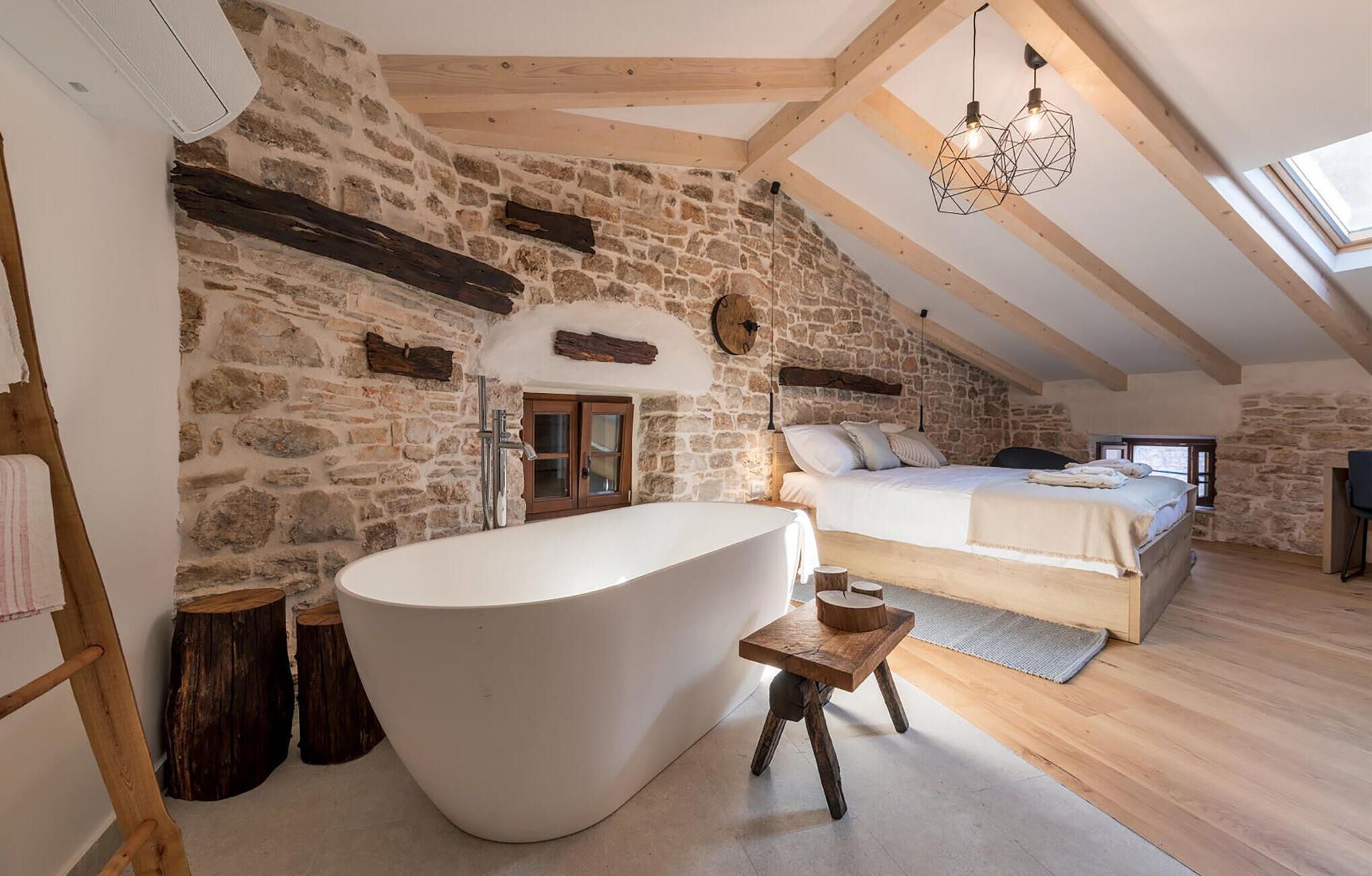 Une chambre avec un mur en pierre, une grande baignoire, un plafond en bois et une lucarne.