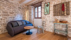 Una stanza accogliente con parete in pietra, soffitto in legno, un divano, un piccolo tavolo rotondo, una sedia e una mensola rustica con una ciotola.