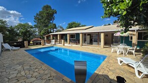 Une piscine aux bords recouverts de pierres, un patio abrité avec des chaises, et une maison avec un toit en tuiles.