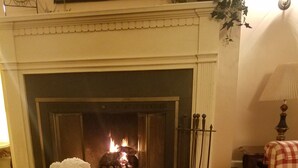 Un salón acogedor con chimenea, un sillón blanco mullido, un sillón a cuadros y una alfombra estampada.