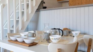Ein Essbereich mit einem weißen Tisch, gedeckt für vier Personen, umgeben von weißen Stühlen. Auf dem Tisch stehen verschiedene Schüsseln, Tassen und eine Kaffeekanne. Im Hintergrund ist eine Holztreppe zu sehen.