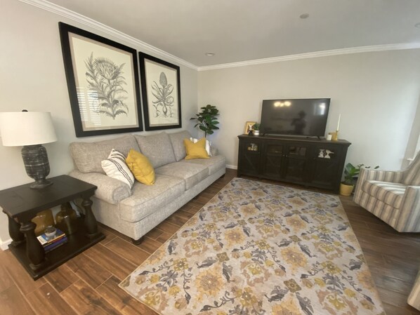 A living room with a grey sofa, a dark wooden coffee table, a patterned rug, a TV on a cabinet, and framed botanical prints on the wall.