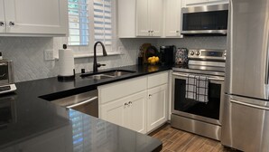 A modern kitchen with white cabinets, stainless steel appliances, a black countertop, and a window with blinds. - Redding Country Cottage (Redding)