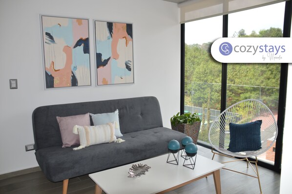 A modern living room with a grey sofa, a white coffee table, and a white chair. There are abstract paintings on the wall and a balcony with a view of trees.