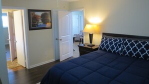 A bedroom with a bed, a ceiling fan, a nightstand with a lamp, and a door leading to another room.