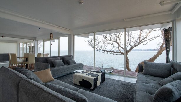 A modern living room with a large window overlooking a body of water, a sofa set, and a dining area.