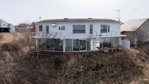 A modern two-story house with a large glass balcony, surrounded by dry vegetation.