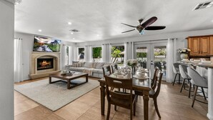 A spacious living room with a fireplace, a dining area with a table set for four, and a kitchen area with wooden cabinets. - Large Home 12 beds Gameroom Heated Salt Pool PPG (Palm Desert)