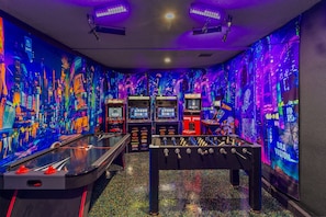 A room with a foosball table, arcade machines, and a neon cityscape mural.