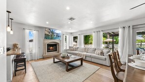 A spacious living room with a fireplace, a sofa set, a dining area, and large windows with curtains. - Large Home 12 beds Gameroom Heated Salt Pool PPG (Palm Desert)