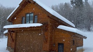Een houten chalet met een grote garage en een met sneeuw bedekte dak, omgeven door bomen.