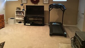 A living room with a TV, bookshelf, treadmill, and a wreath on the wall. - Enjoy Northern MN (Cook)