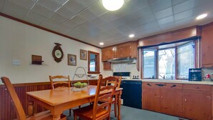 A dining area with a wooden table and chairs, a kitchen with wooden cabinets, a dishwasher, and a window with blinds. - Huge historic on Lake Michigan w/ large yard, fire pit, BBQ & air conditioning (Manistee)