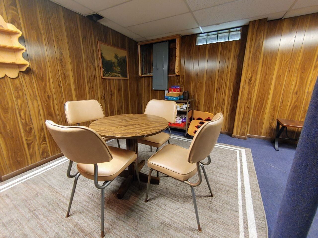 A room with a wooden paneled wall, a round wooden table with four beige chairs, a small bookshelf, and a window with a view of trees.