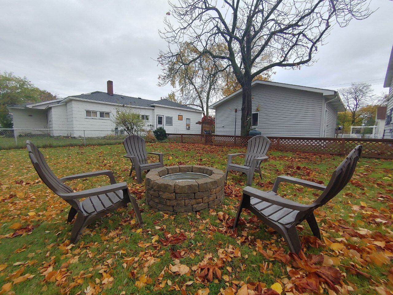 A backyard with a fire pit, four Adirondack chairs, and a large tree.